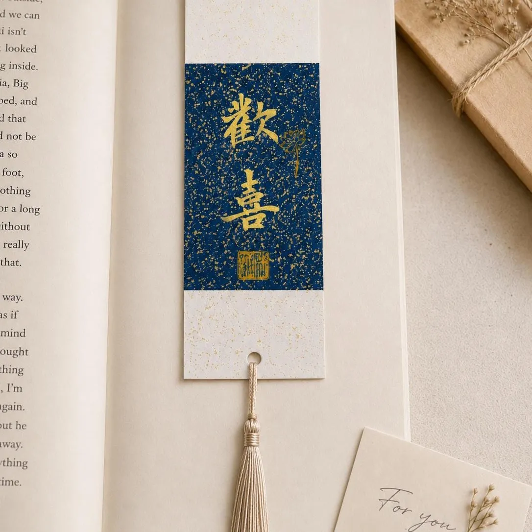 A calligraphy bookmark on indigo speckled paper with a cream tassel, resting in an open book beside a wrapped gift and a "For you" card