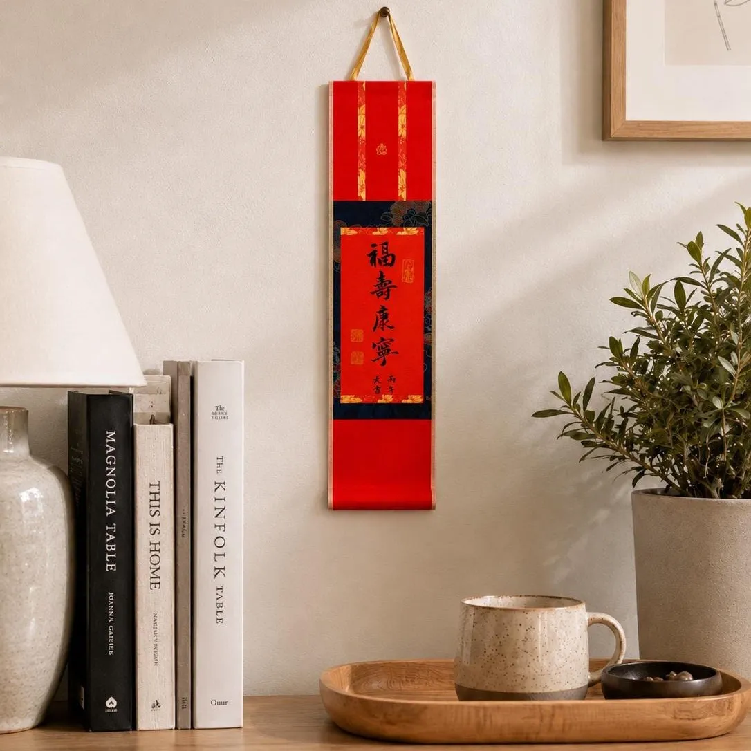 A red mini calligraphy wall scroll with a blue brocade panel, hanging above a wooden sideboard beside books, a potted olive branch and a stoneware mug